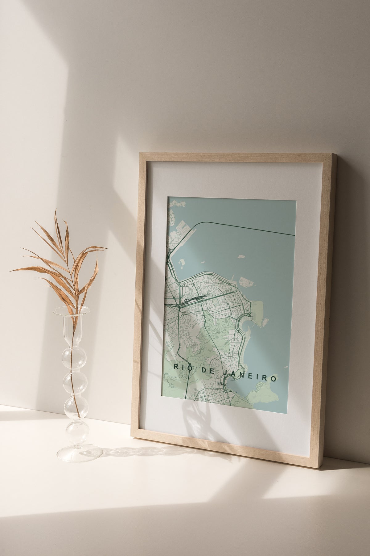 Rio de Janeiro, Brazil map print in the Forest theme in a light wooden frame on a shelf beside a glass vase