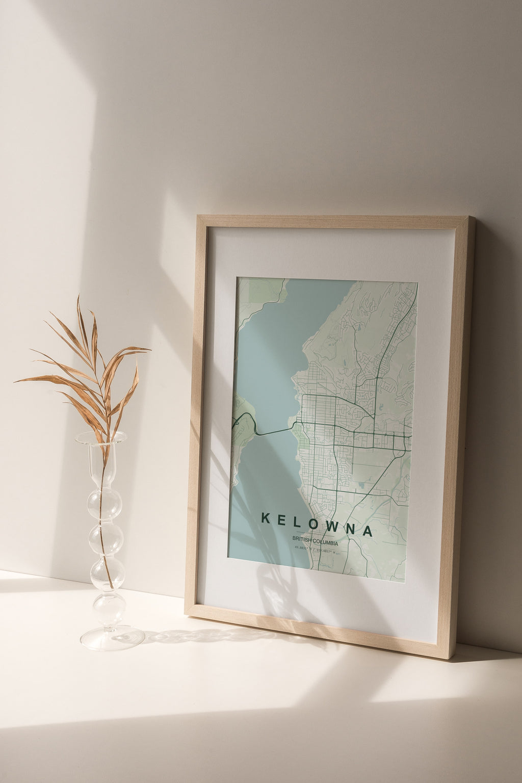 Kelowna, British Columbia, Canada map print in the Forest theme in a light wooden frame on a shelf beside a glass vase