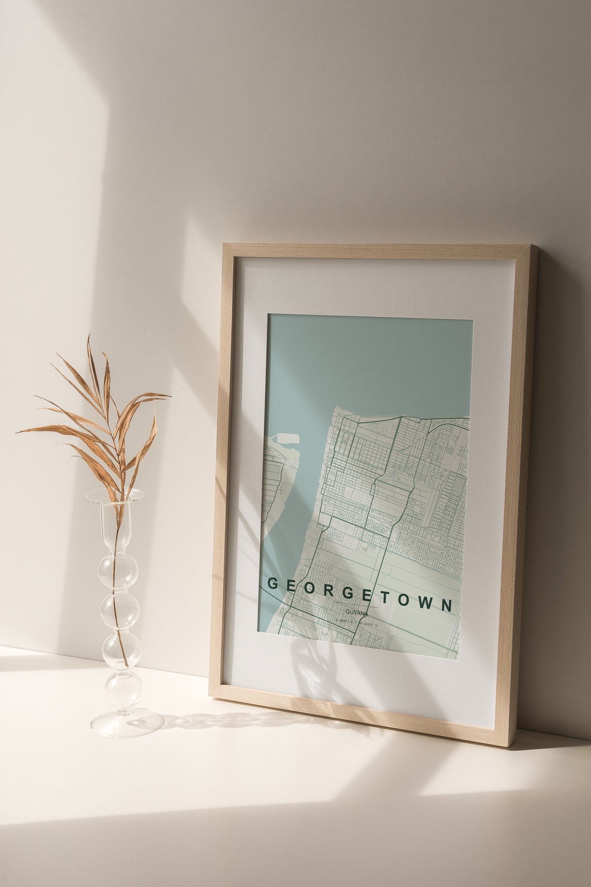 Georgetown, Guyana map print in the Forest theme in a light wooden frame on a shelf beside a glass vase