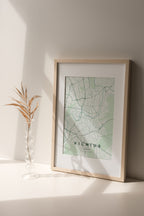 Vilnius map print in the Forest theme in a light wooden frame on a shelf beside a glass vase