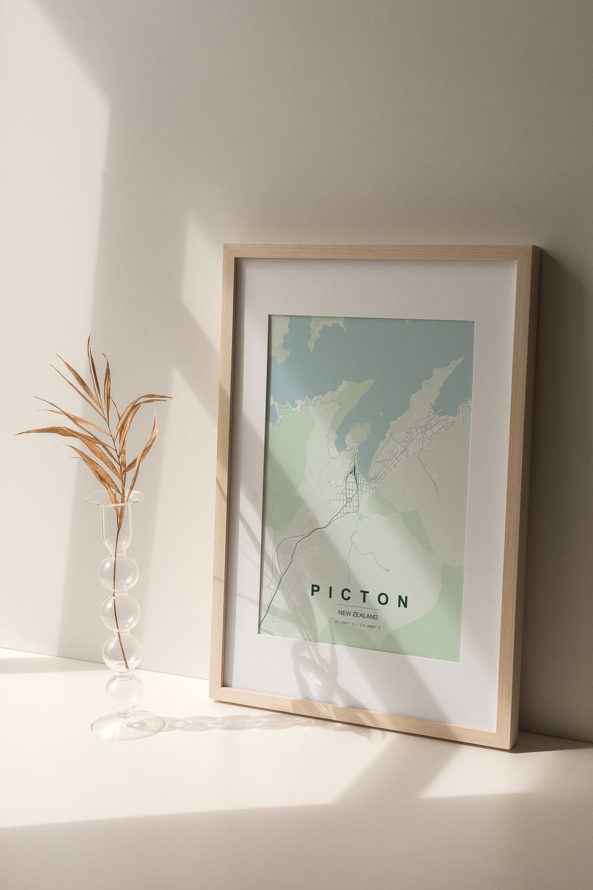 Picton, Marlborough, New Zealand map print in the Forest theme in a light wooden frame on a shelf beside a glass vase