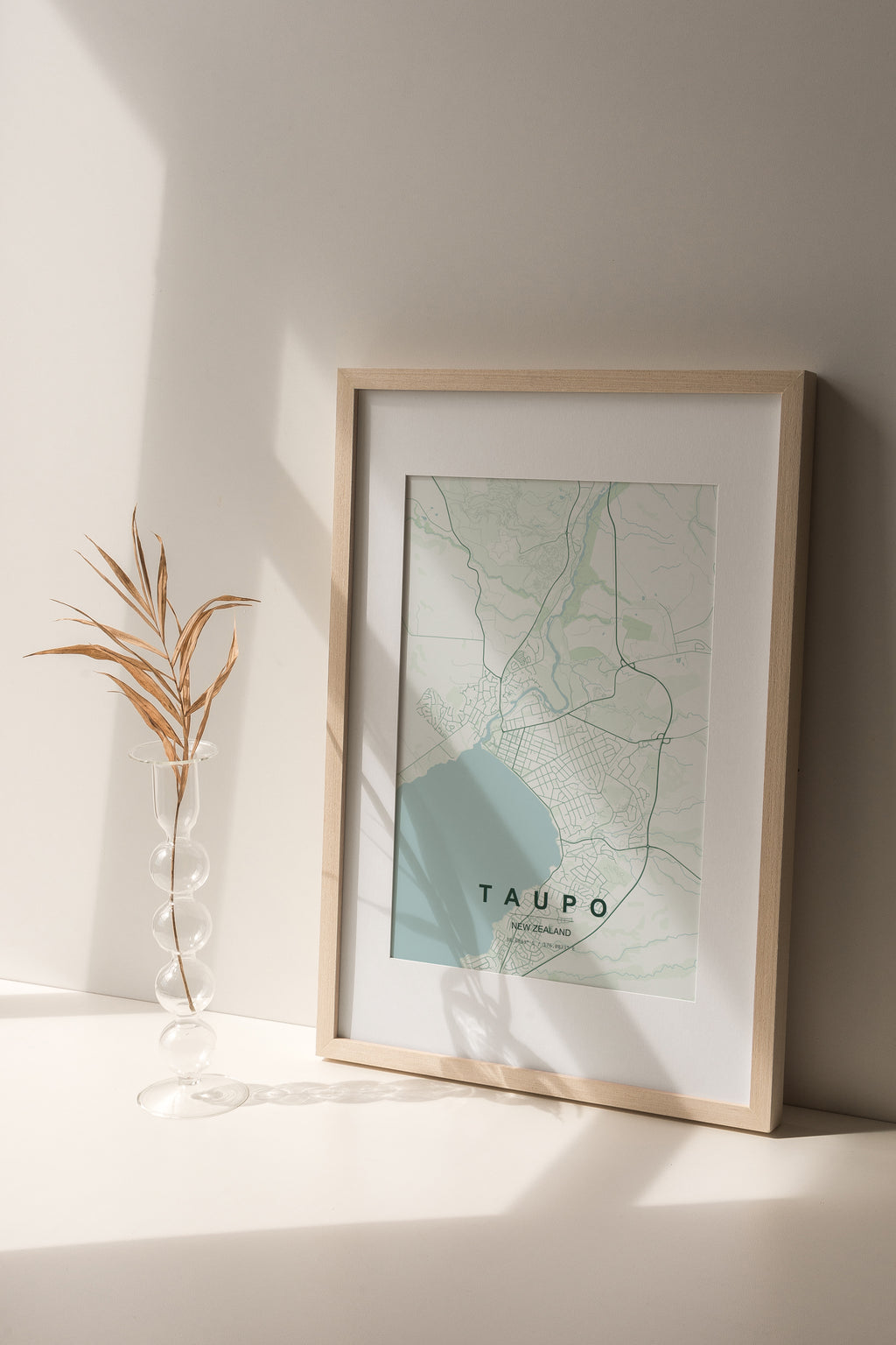 Taupō, New Zealand map print in the Forest theme in a light wooden frame on a shelf beside a glass vase