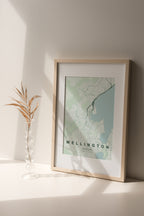 Wellington, New Zealand map print in the Forest theme in a light wooden frame on a shelf beside a glass vase