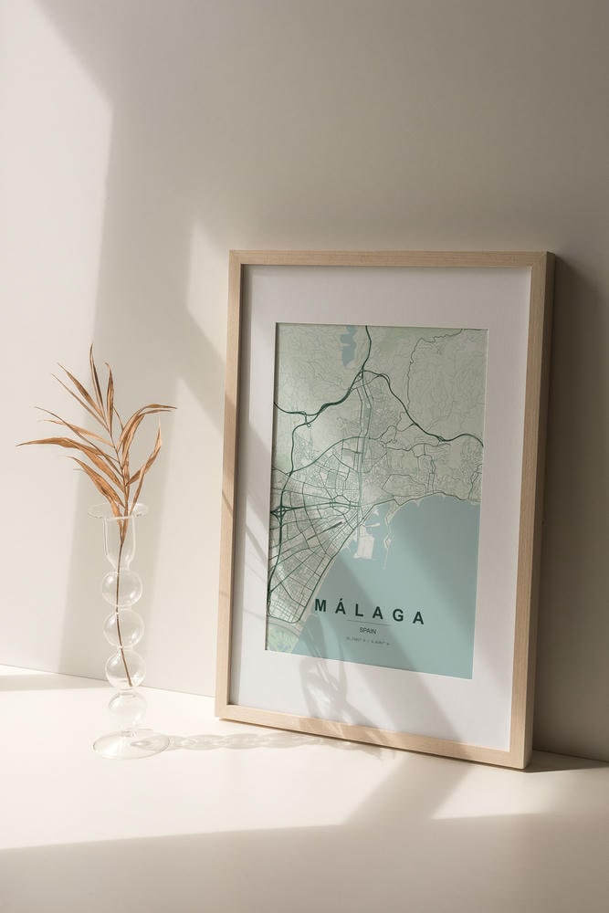 Málaga map print in the Forest theme in a light wooden frame on a shelf beside a glass vase
